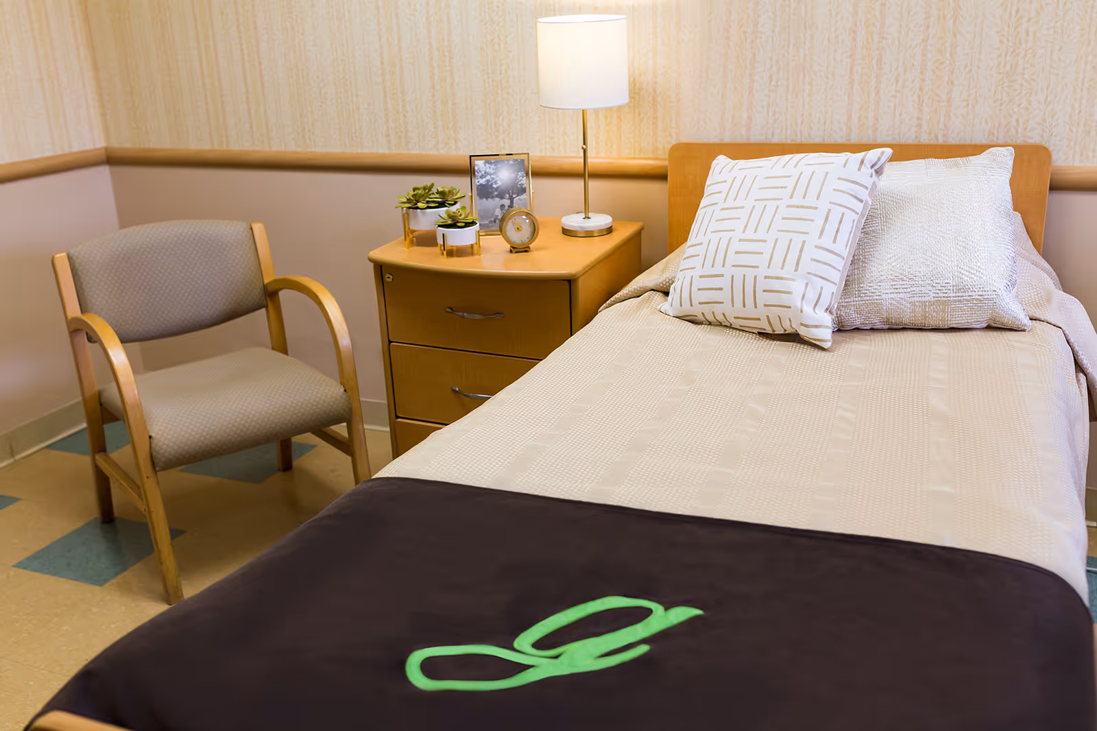 A neatly made single bed with two decorative pillows and a dark blanket with a green letter 'g' embroidered on it. Next to the bed is a wooden nightstand with a lamp, a small clock, a framed photo, and two small potted plants. A wooden armchair with a cushioned seat and backrest is positioned beside the nightstand. The room has beige wallpaper and a tiled floor with a pattern of beige and blue squares.