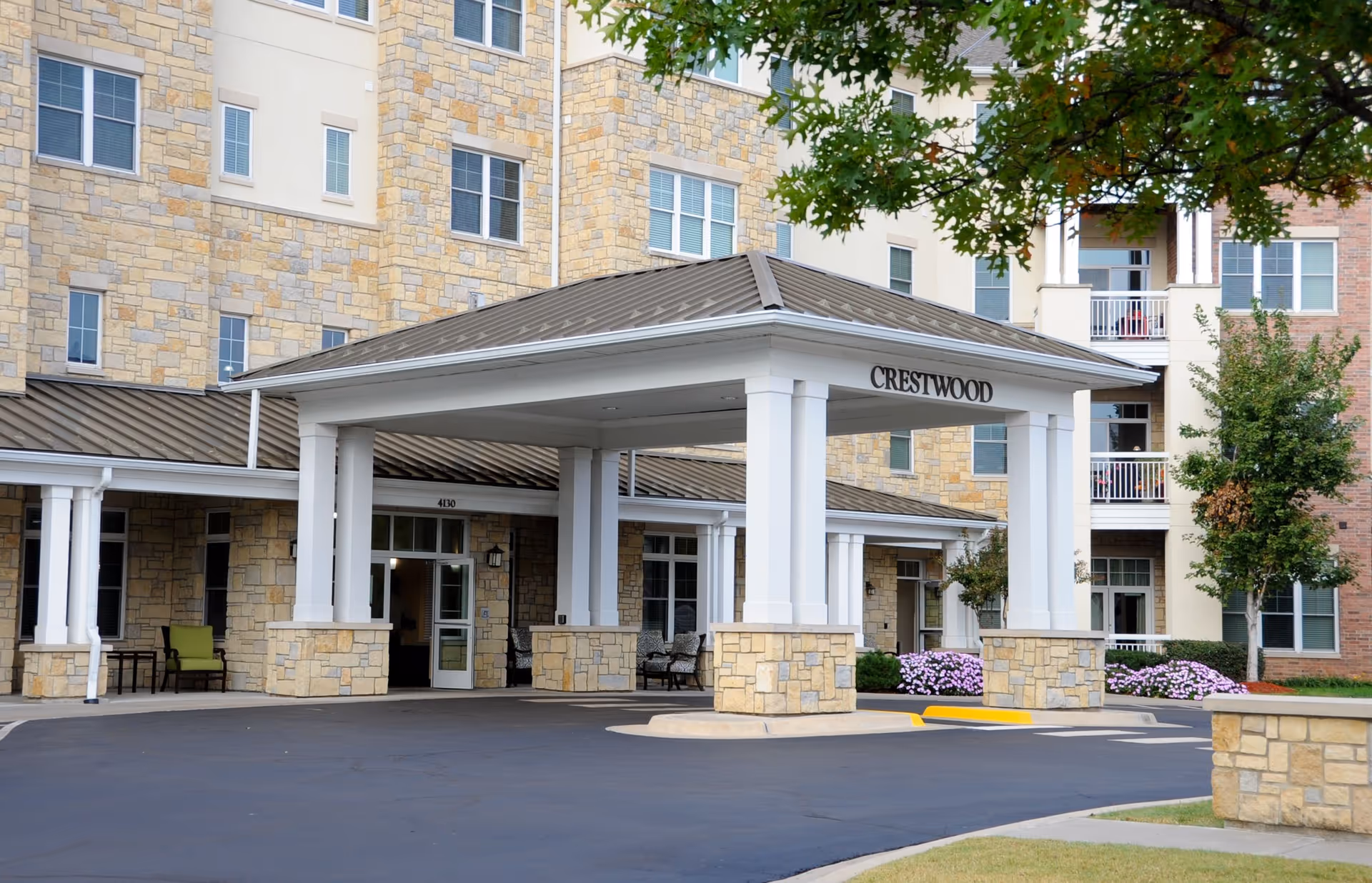 Entrance of a senior living facility named Crestwood with a covered drop-off area supported by white columns and stone bases. The building exterior features a mix of stone and brick with multiple windows and balconies. There are some green chairs and plants near the entrance, and a tree with green leaves is visible on the right side.