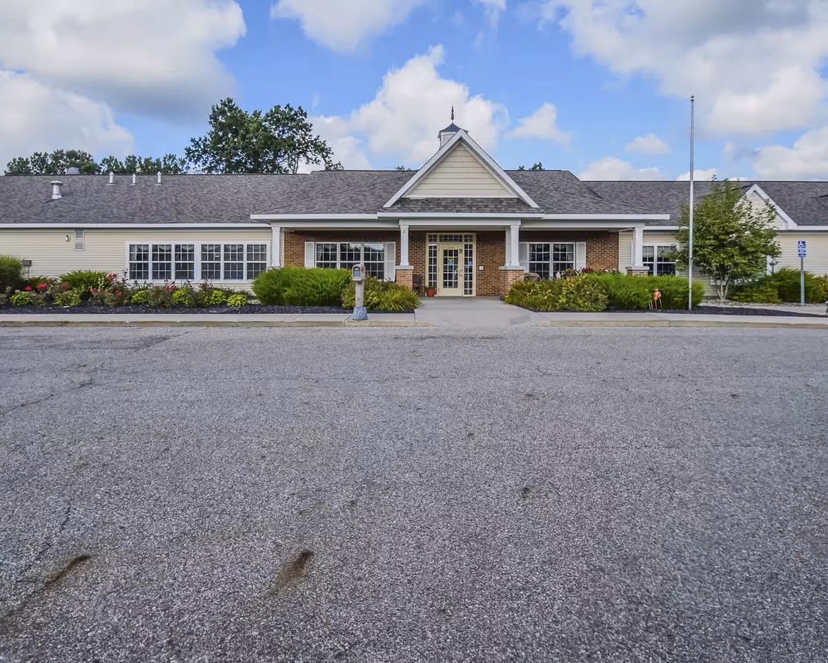 Single-story assisted living facility with brick entrance, manicured shrubs, and a wide parking area.