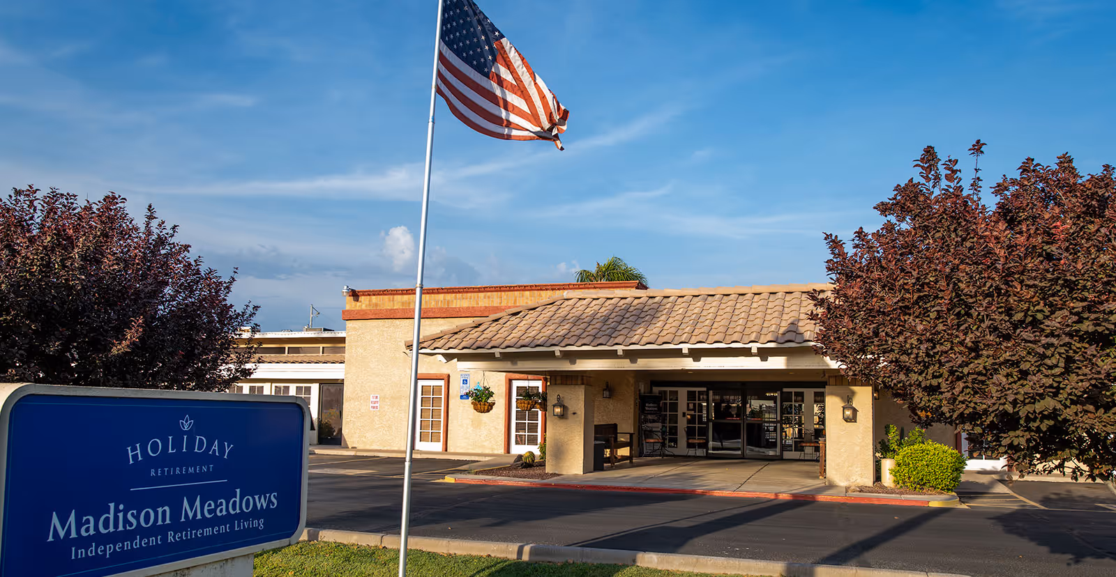 Front entrance and facade of Madison Meadows Retirement Community with an American flag and facility sign in the foreground.