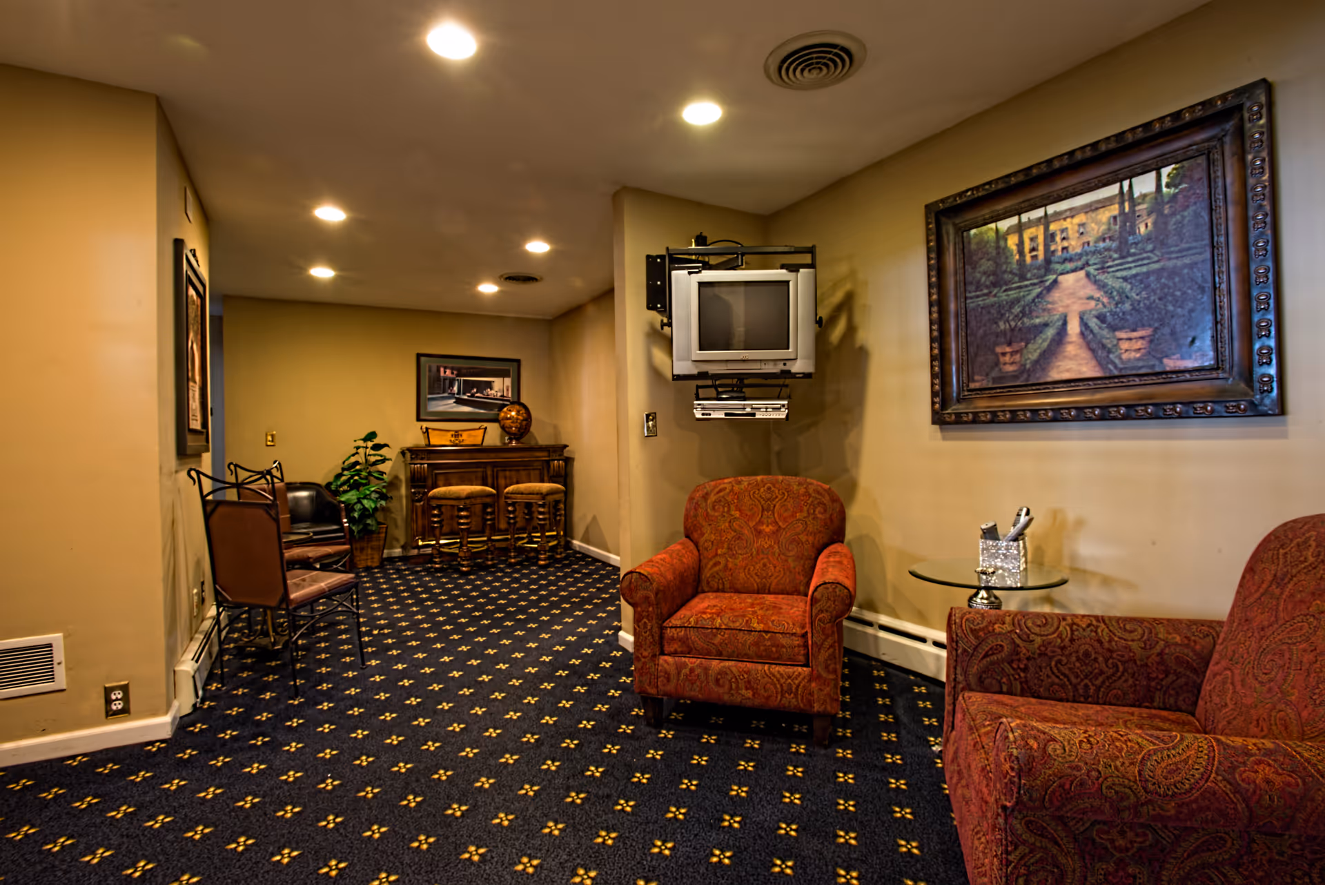 A cozy senior living lounge with patterned carpet, red upholstered chairs, and wall art.