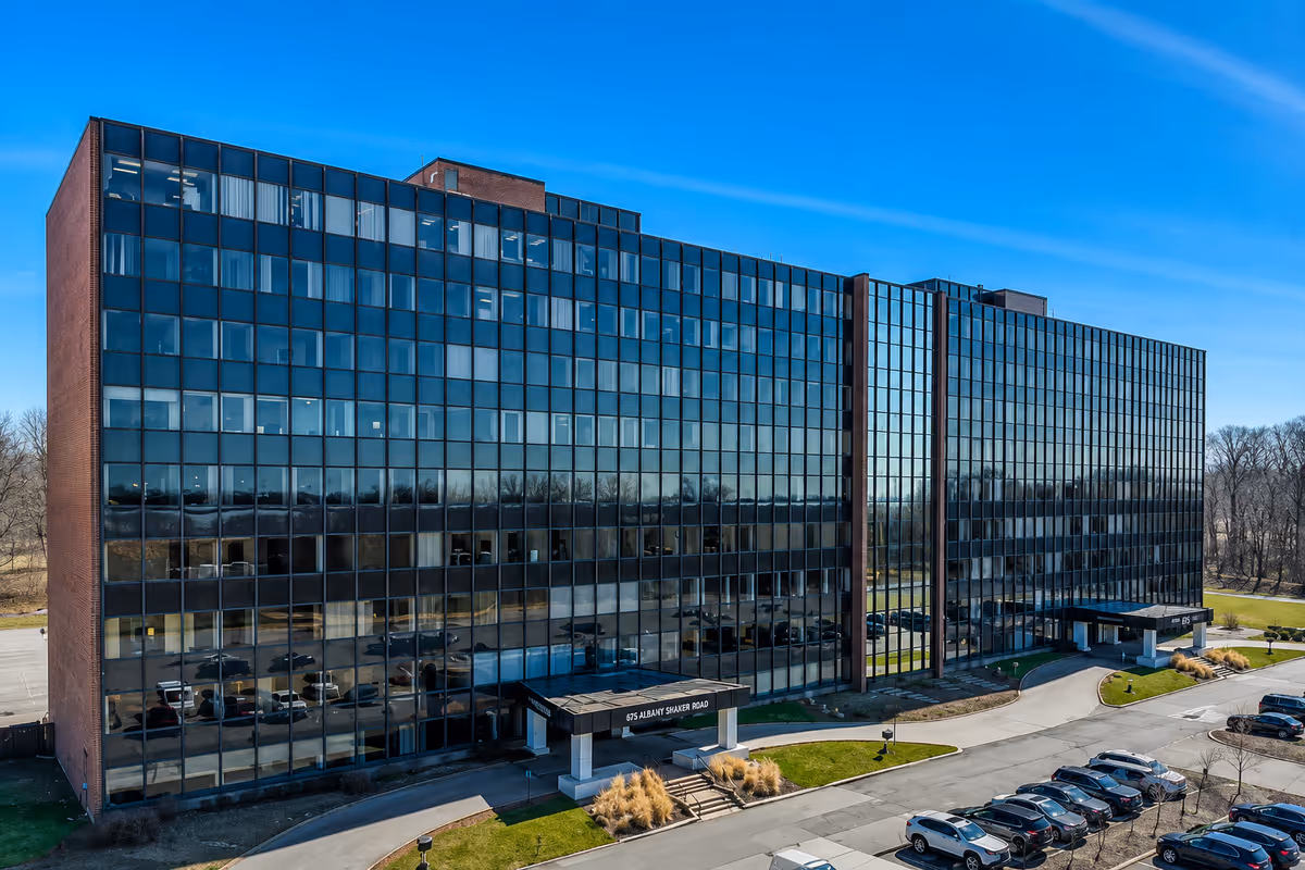 A modern glass-front office building with a parking lot and clear blue sky.