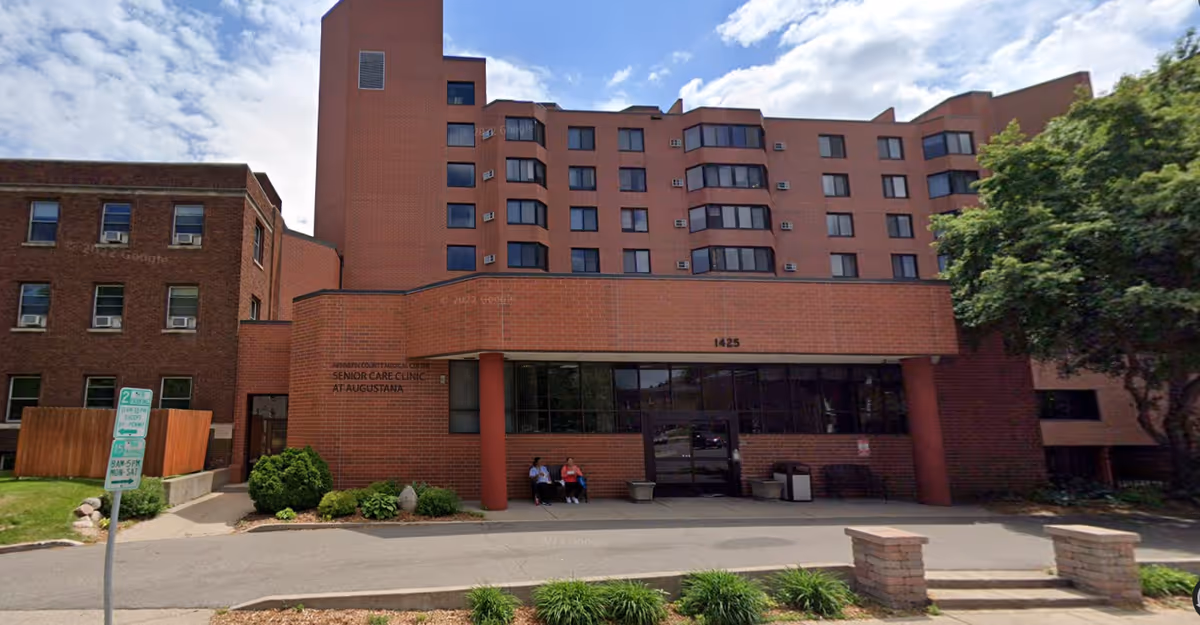 Brick senior living building exterior with entrance and a few residents seated outside.