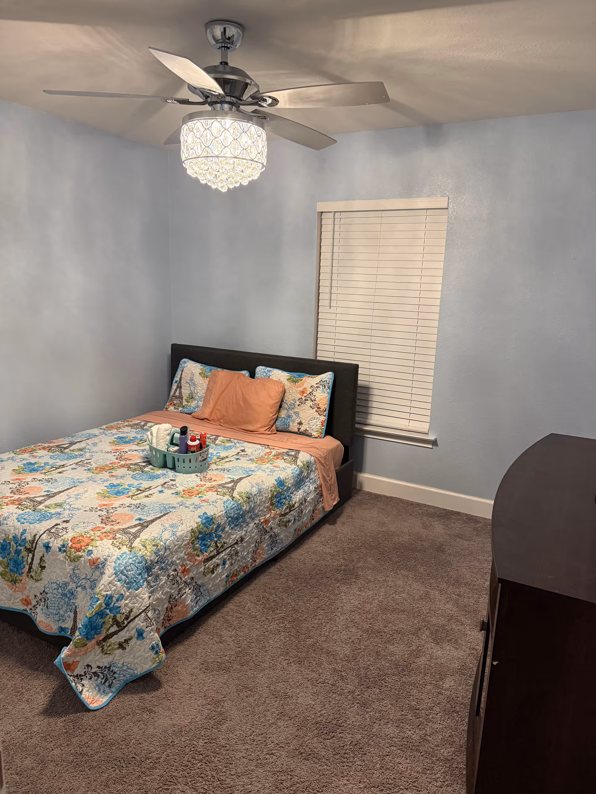 A blue-painted bedroom with a floral quilted bed, window blinds, and a ceiling fan.