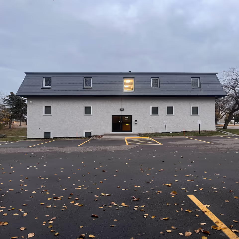 Two-story white brick building with a dark slate roof and a front parking lot.