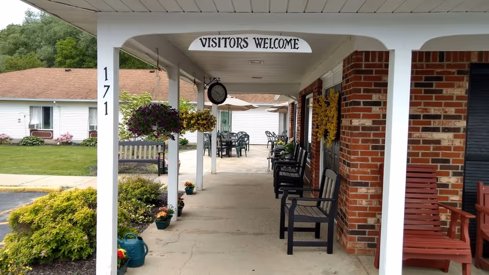 Covered front walkway of The Cloisters with a 'VISITORS WELCOME' sign, chairs, hanging plants, and a brick facade.