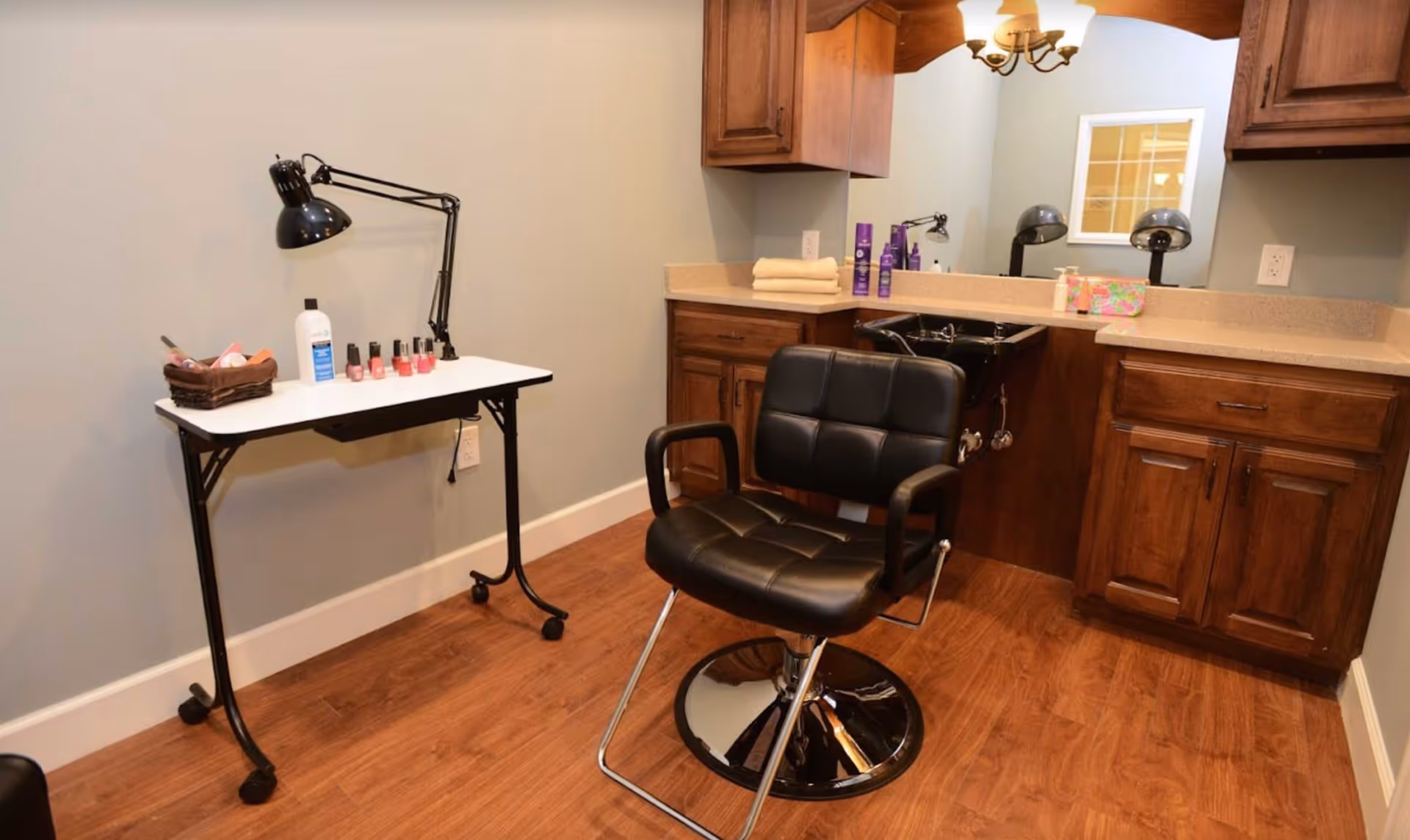 A small salon or grooming area with a black salon chair in front of a wooden cabinet and sink. A white table with nail polish bottles, a lamp, and other manicure supplies is positioned against a light blue wall. The floor is wooden.