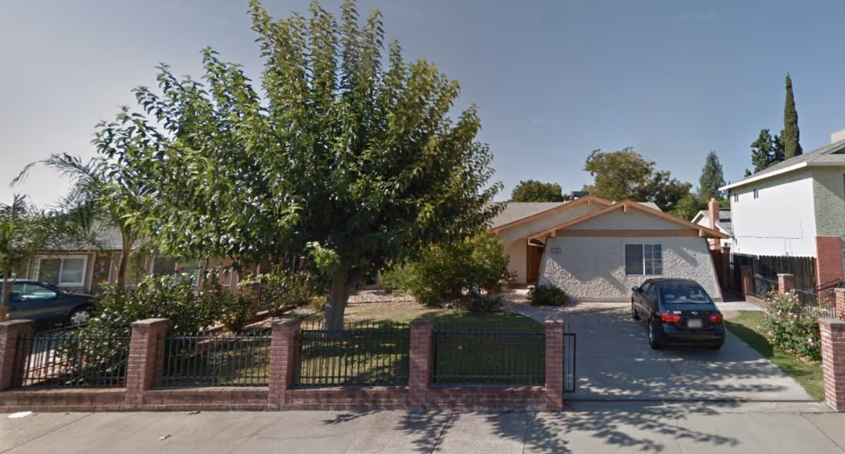 Single-story house with a front yard, large tree, brick fence, and a car parked in the driveway.