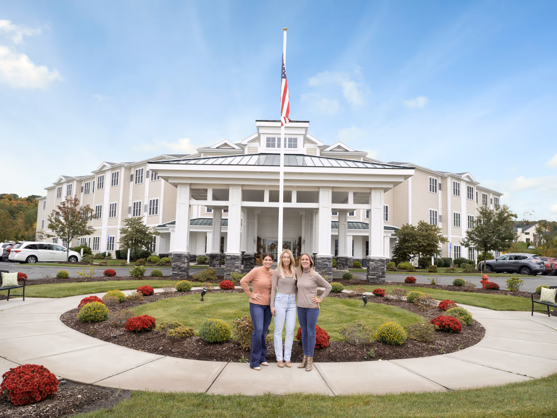 Three women standing together smiling in front of a large, modern assisted living facility building with a covered entrance and an American flag on a flagpole. The area around them is landscaped with green grass, red and yellow flowers, and a curved concrete walkway.