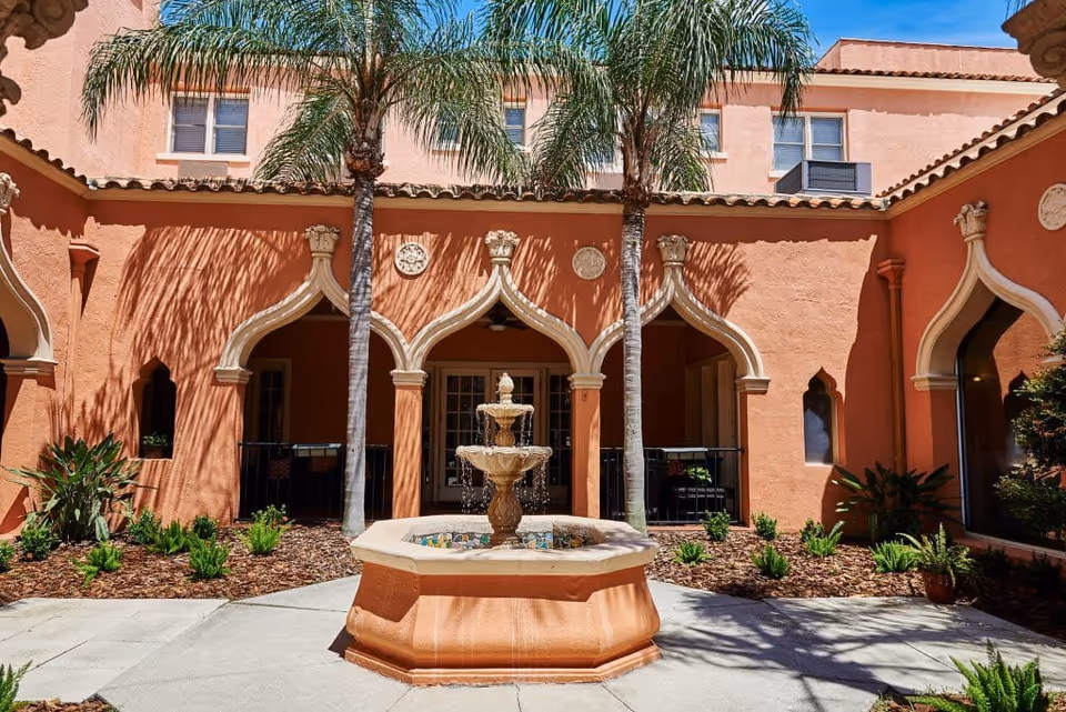 Outdoor courtyard area with a central multi-tiered water fountain surrounded by plants and palm trees. The courtyard is enclosed by a building with peach-colored walls and arched doorways and windows, featuring decorative architectural details.