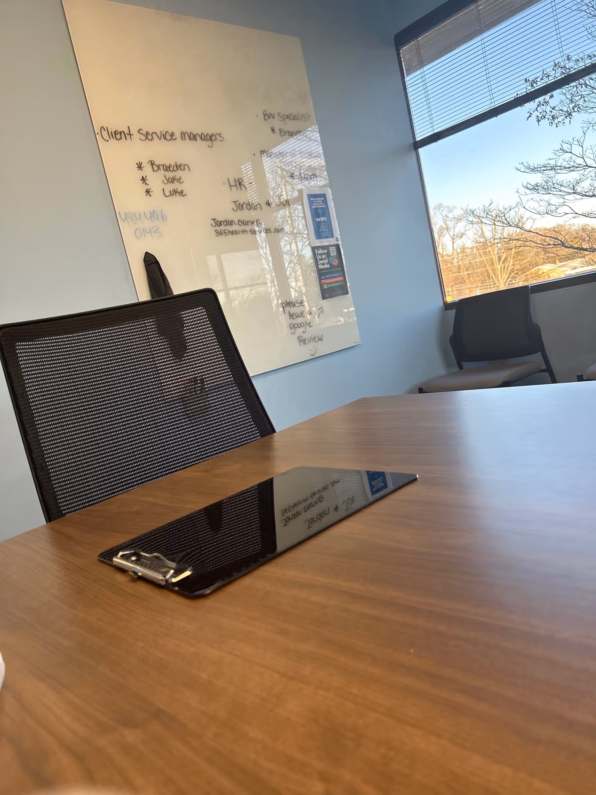 A conference room with wooden table, mesh chair, whiteboard, window blinds, and a tablet.