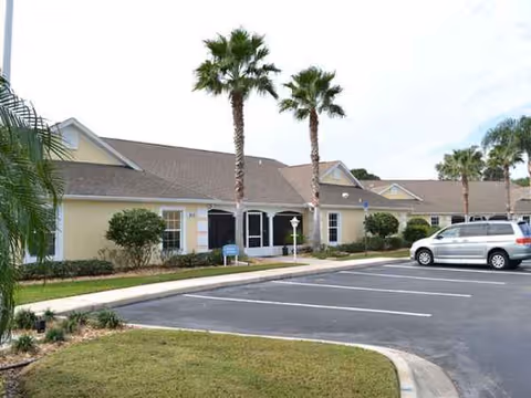 Single-story senior living facility with palm trees, landscaped lawn, and a parking lot in front.
