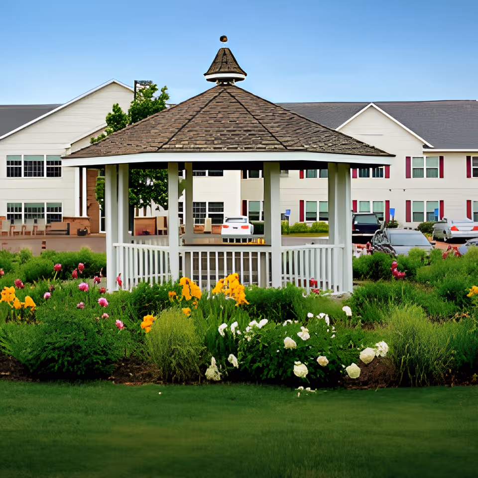 A white gazebo with a shingled roof is surrounded by colorful flowers and green shrubs in a garden area. Behind the gazebo, there is a large building with white siding and multiple windows, along with several parked cars. The sky is clear and blue.