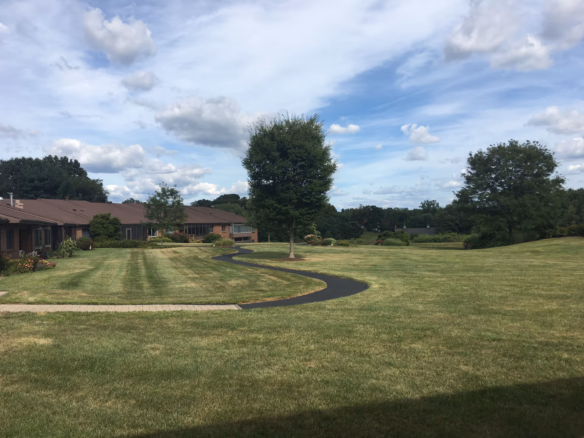 Curved asphalt path winds through a manicured lawn beside low-rise facility buildings.