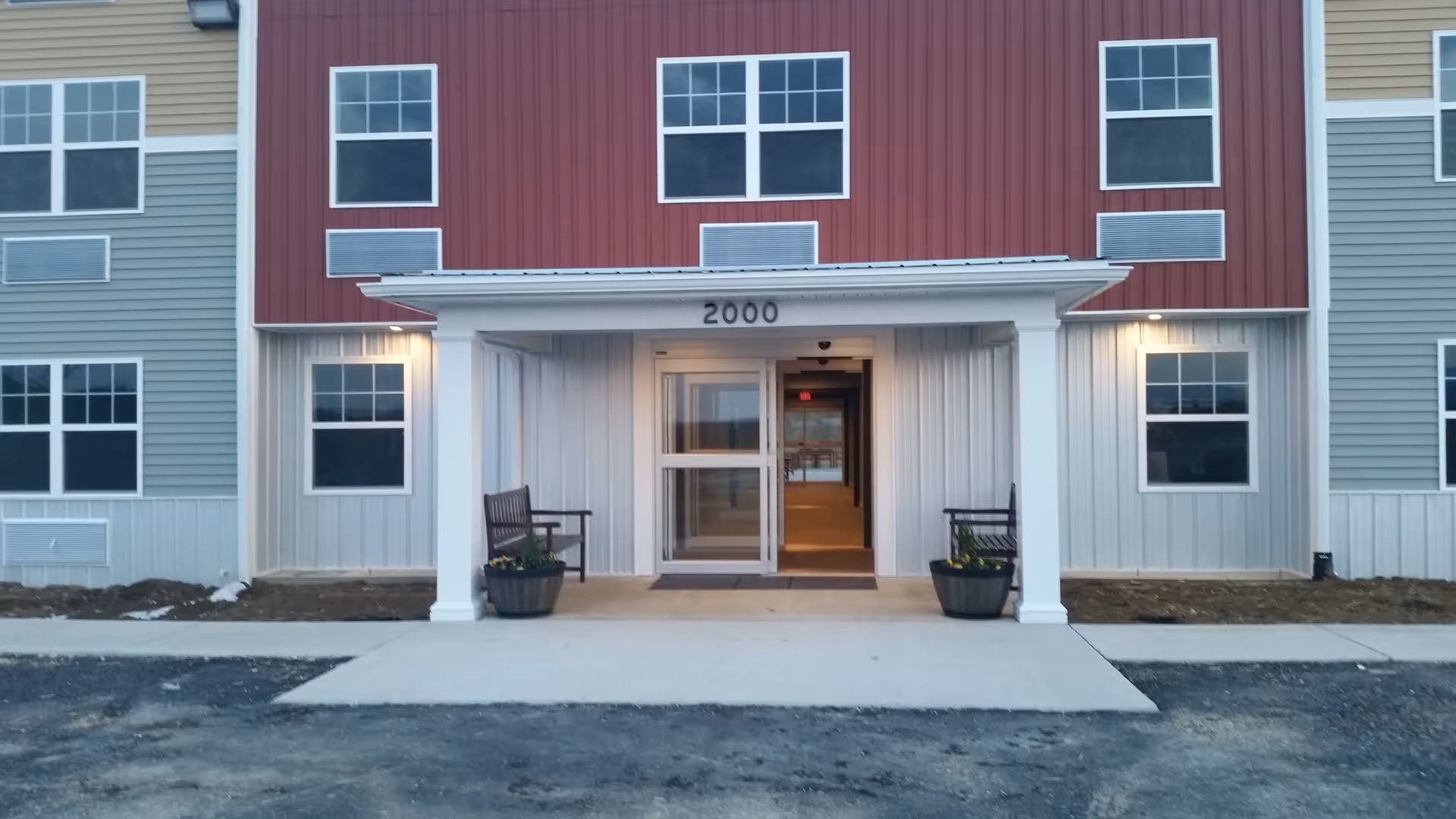 Entrance of a senior living facility named The View, showing a covered porch with two benches and potted plants on either side of the doorway. The building exterior features red and gray siding with multiple windows and the number 2000 above the entrance.