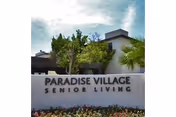 A sign reading Paradise Village Senior Living sits before a building with palm trees.