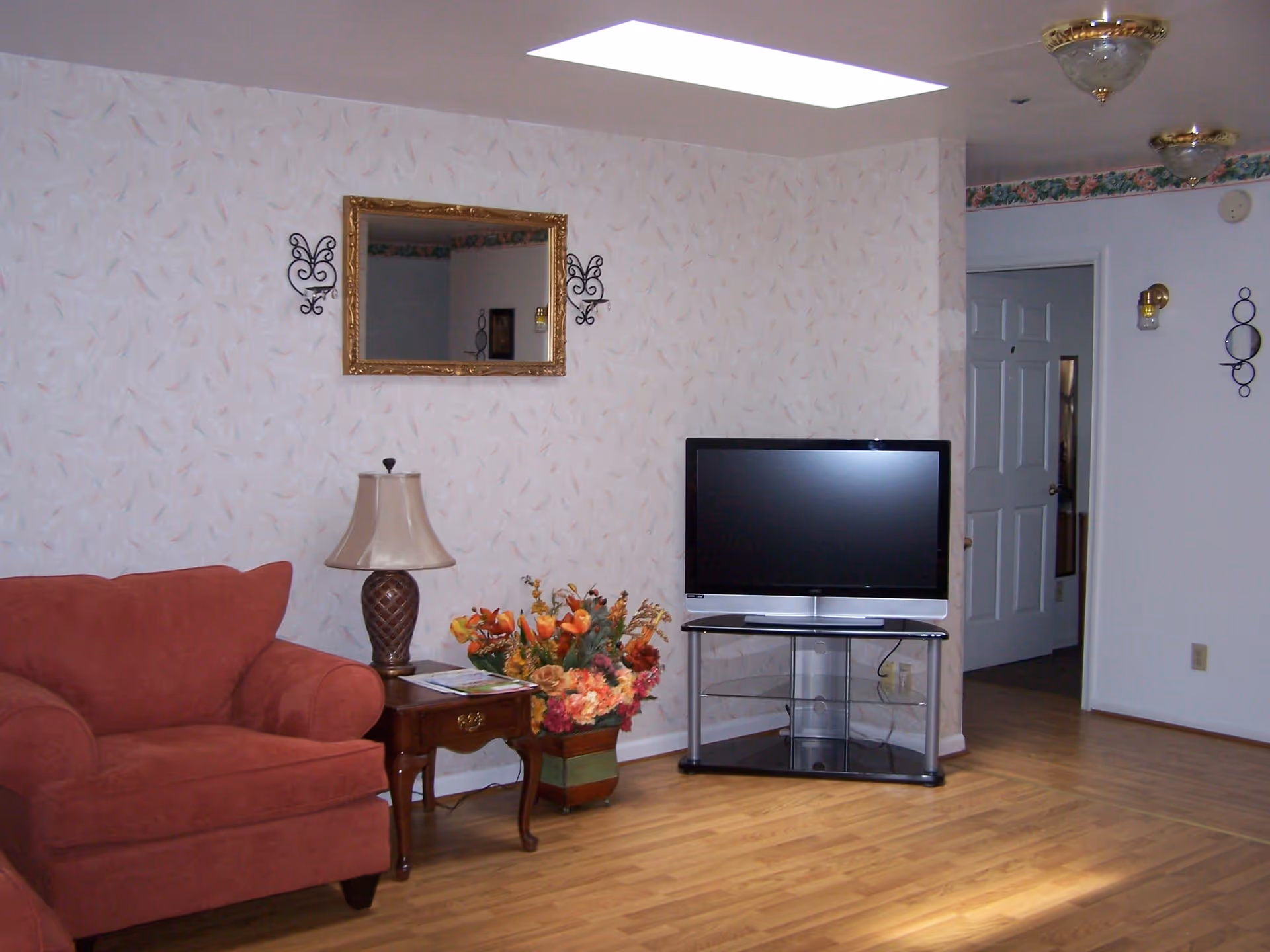 A living room with a red upholstered armchair, a wooden side table with a lamp and a floral arrangement, a flat-screen TV on a glass stand, and a wall mirror with decorative sconces on either side. The room has light-colored wallpaper with a subtle pattern and wood flooring.