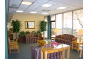 Cozy senior living lounge with striped sofas, wooden tables, plants, and large windows.