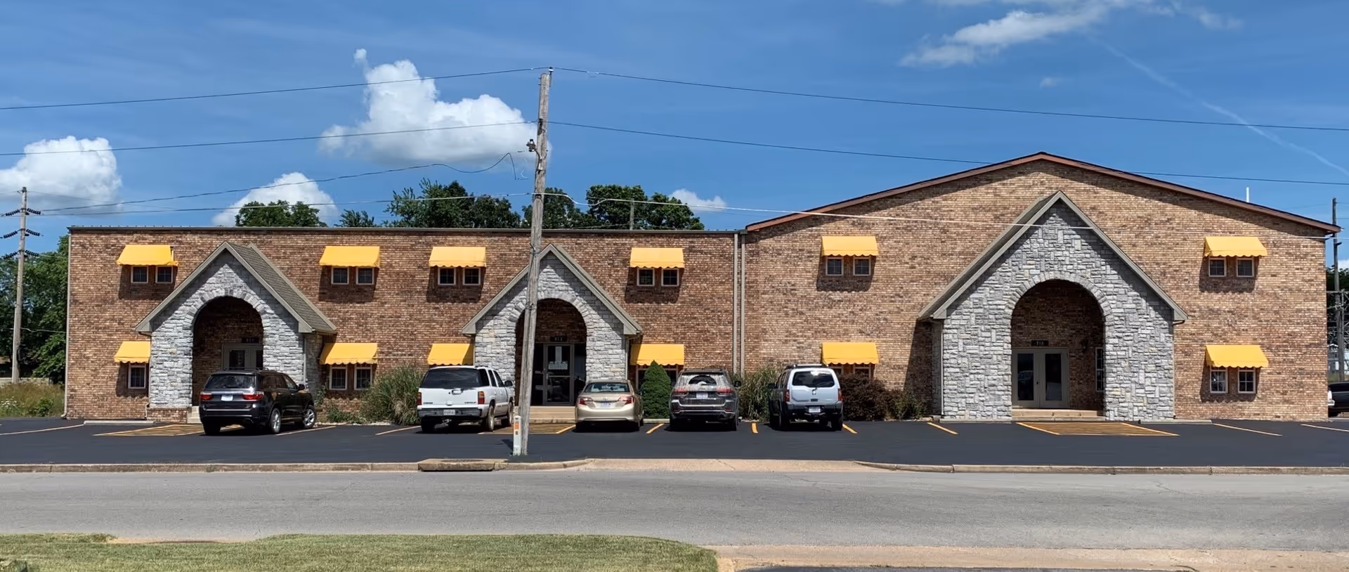 Brick facility with arched entrances, yellow awnings, and parked cars in a strip along the front.