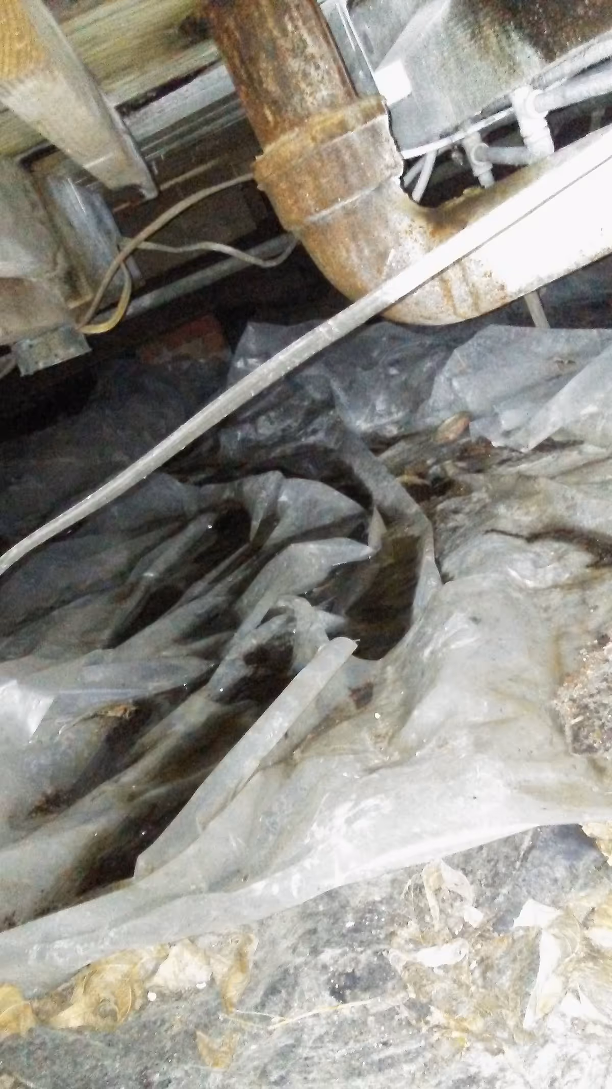 Rusty pipes and plastic sheeting clutter a dirty interior under a sink.