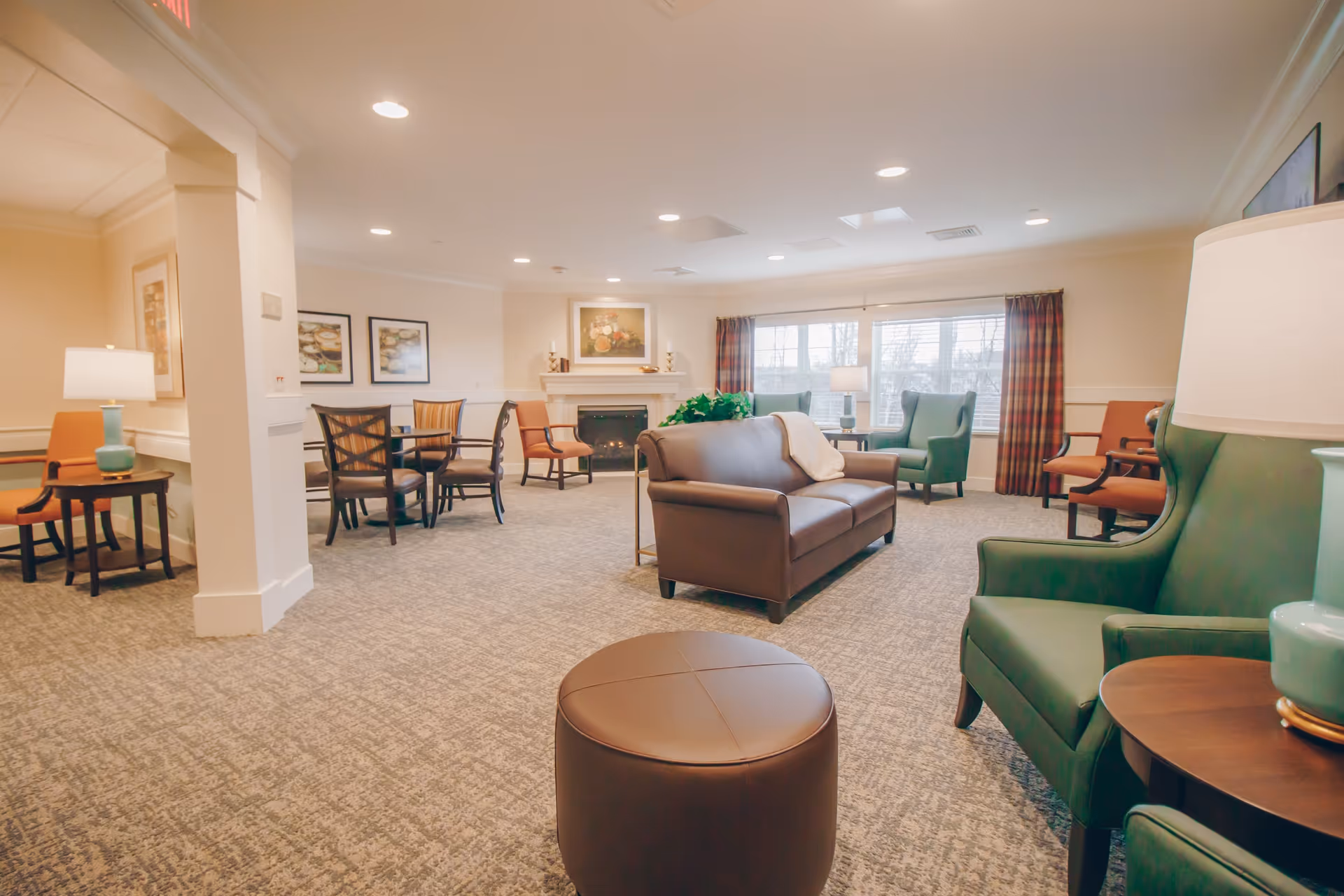 A spacious senior living common area with a brown leather sofa, green armchairs, and orange chairs arranged around small tables. There is a fireplace with a floral painting above it, large windows with plaid curtains, and soft lighting from ceiling lights and table lamps.