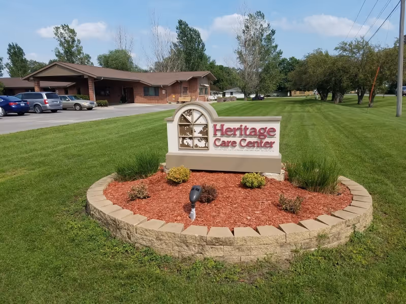 Outdoor view of Heritage Care Center sign surrounded by a circular flower bed with mulch and small plants, with a building and parked cars in the background on a sunny day.