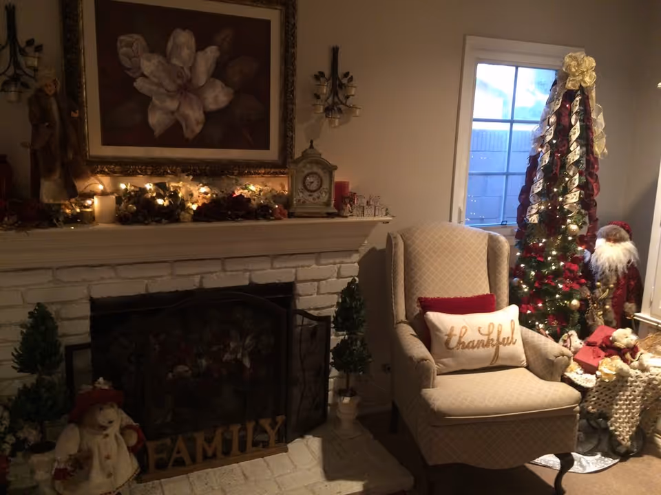 Cozy living room with a decorated fireplace, an upholstered armchair with a "thankful" pillow, and a decorated Christmas tree.