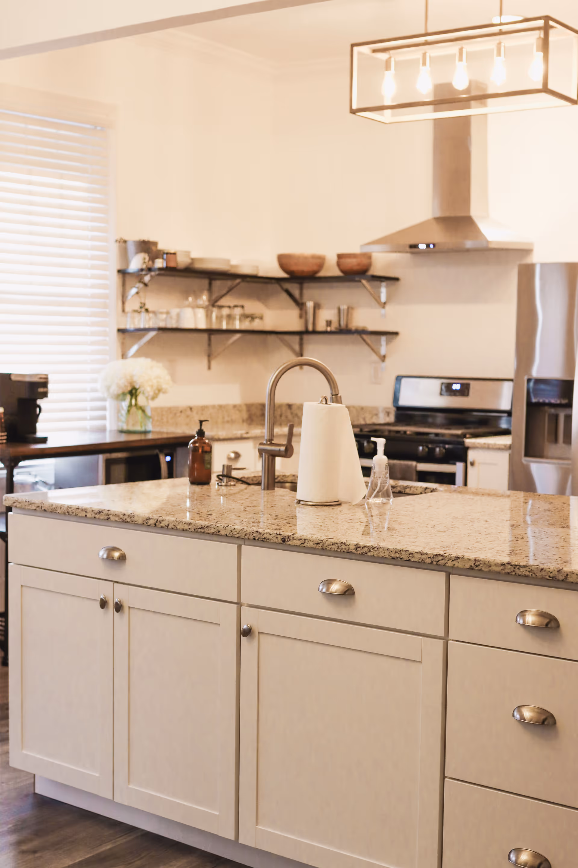 Bright modern kitchen with granite island, sink, open shelving, and stainless steel appliances.