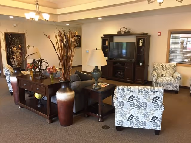 A cozy living room area in an assisted living facility featuring a dark wooden entertainment center with a TV, two patterned armchairs, a gray sofa, a wooden console table with decorative vases and flowers, and a side table with a lamp. The room has beige walls, carpeted floor, and a window with wooden blinds.