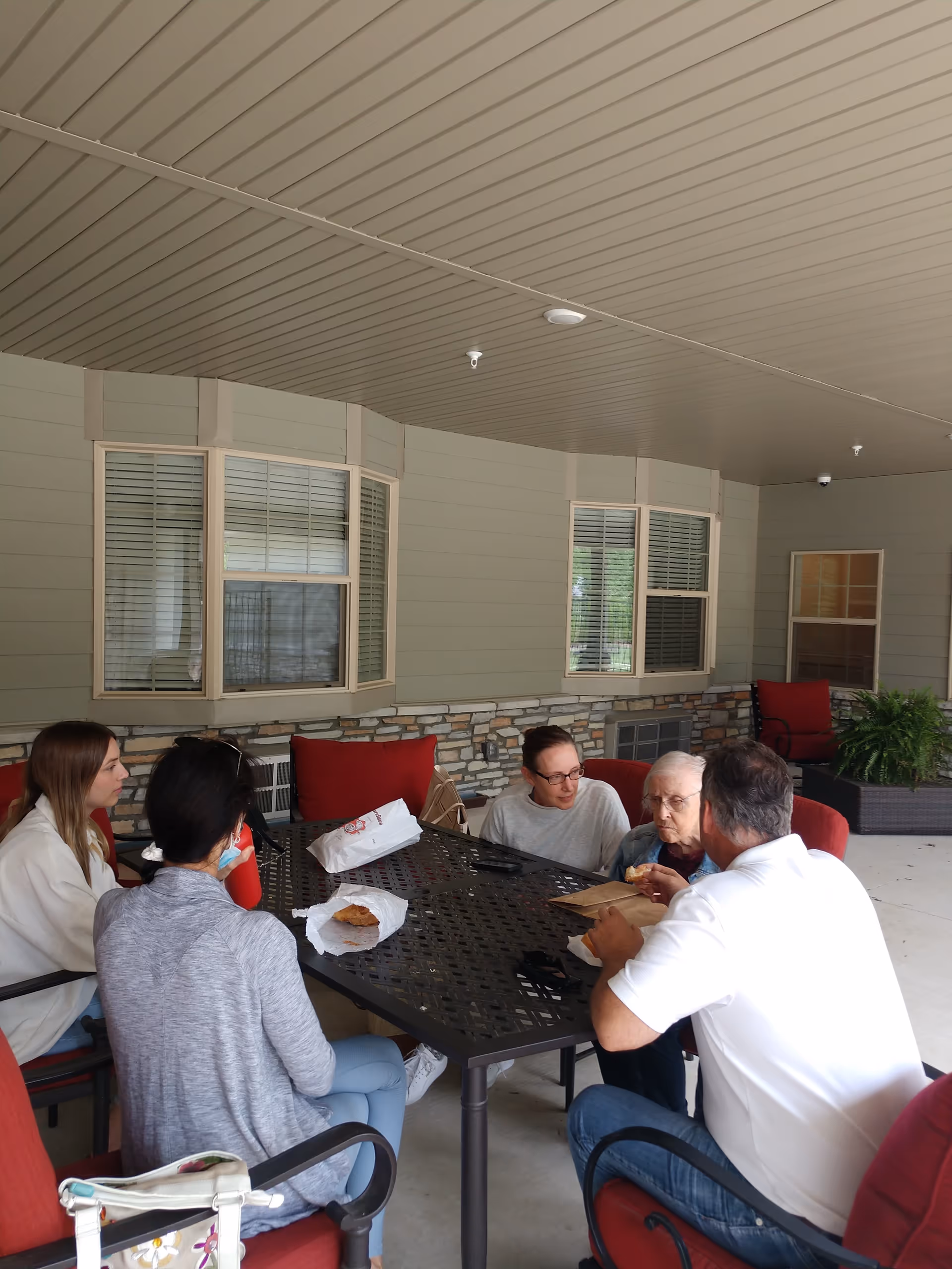 Residents and a caregiver sit around an outdoor table on a covered patio, eating together.