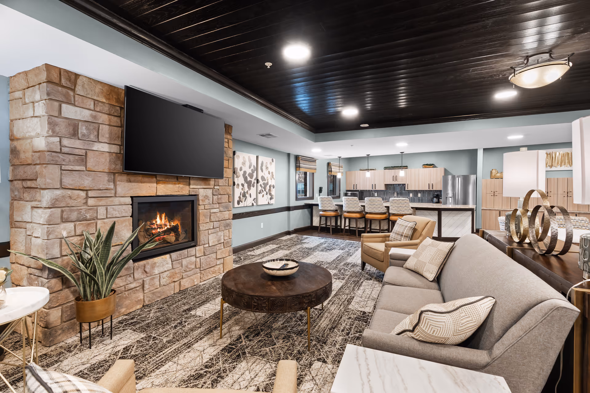 Lounge area with stone fireplace, mounted TV, and kitchen in the background.
