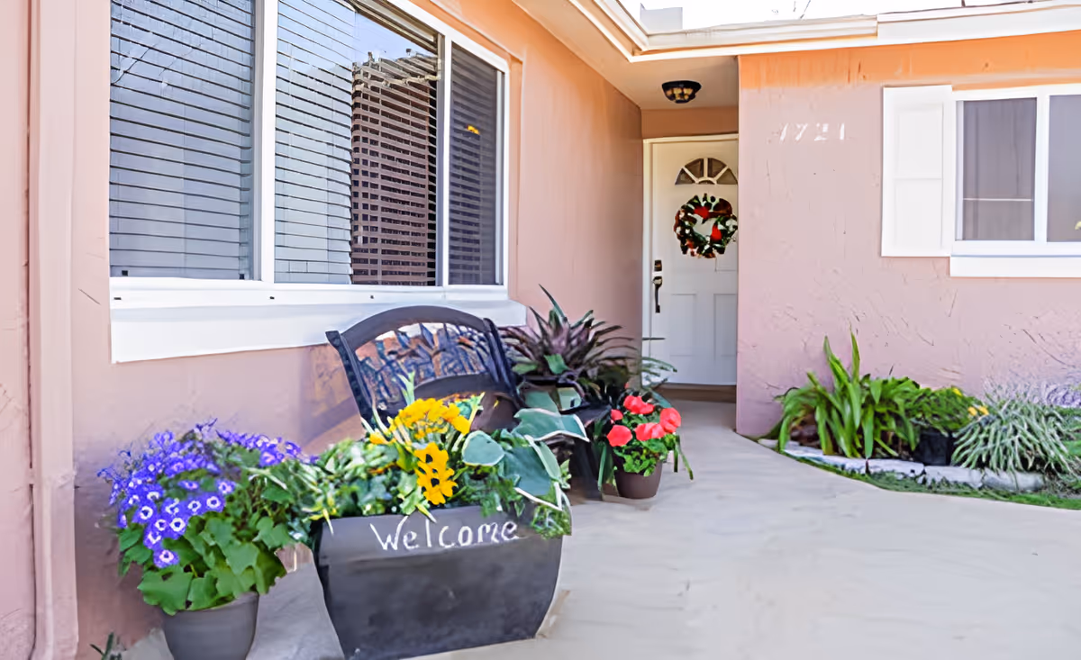 Front entrance of a house with a white door decorated with a wreath. There is a bench with a planter in front of it containing colorful flowers and a 'Welcome' sign. Additional potted plants and greenery are placed along the walkway leading to the door.