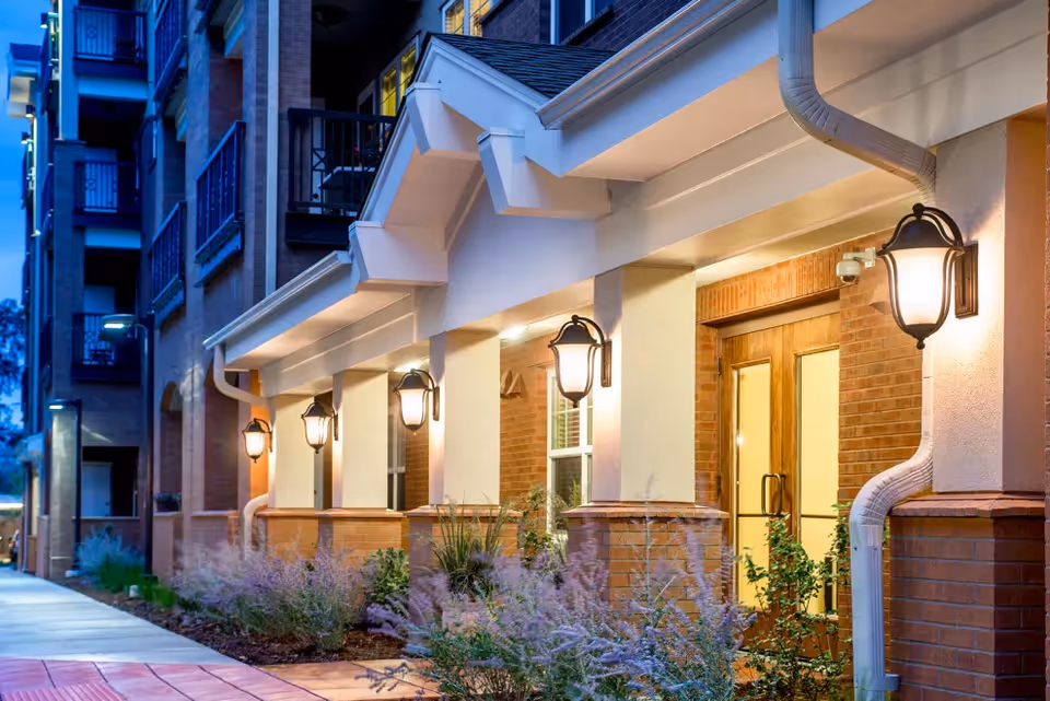 Exterior view of a building entrance at dusk with warm lighting, brick walls, white columns, and decorative outdoor lanterns along a walkway bordered by plants and shrubs.