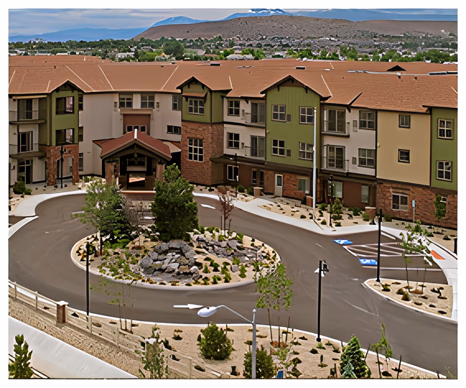 Front entrance and circular driveway of a multi-story senior living building with a landscaped center island and mountains in the background.