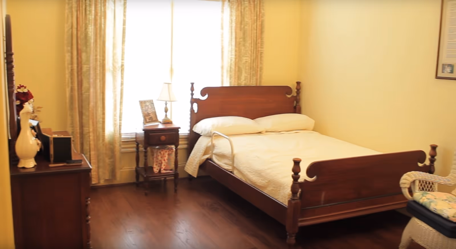 A warmly lit bedroom with a wooden bed, nightstand, dresser and a window with curtains.
