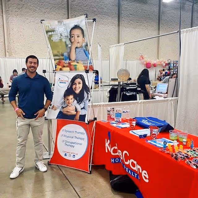 A man stands beside a KidsCare Home Health booth at an indoor expo.