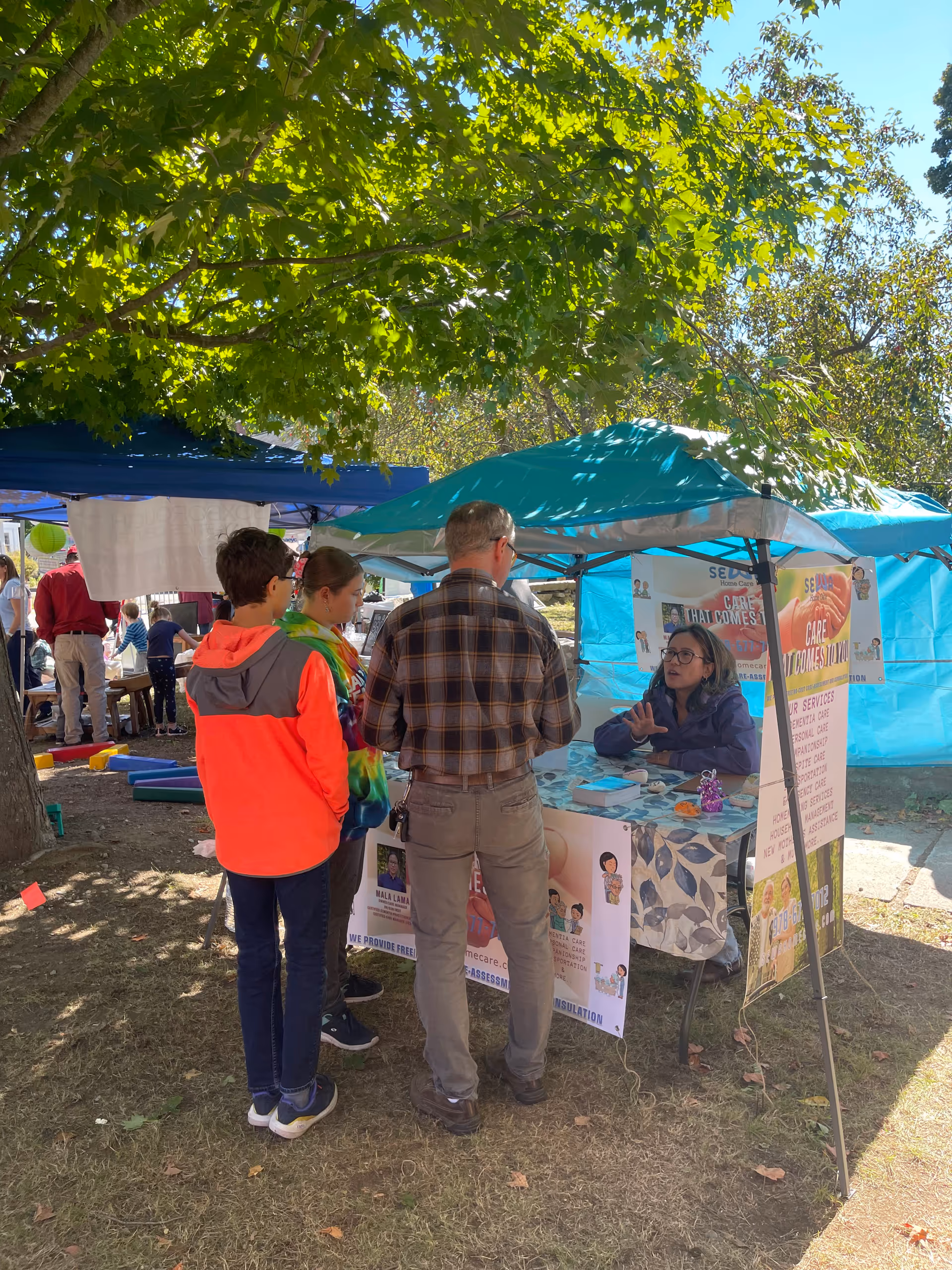 Outdoor SEWA HOME CARE booth under trees, staff talking to visitors at a community fair.