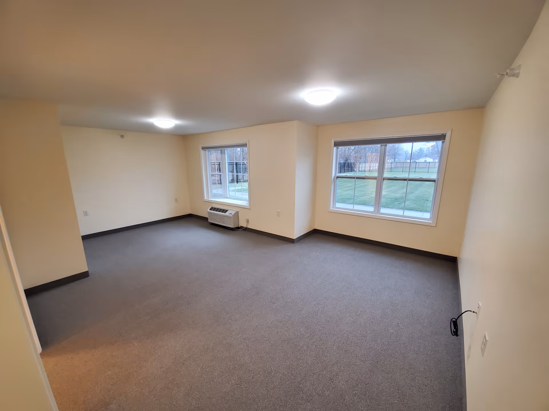 Empty room with beige walls and gray carpet, two large windows showing a grassy outdoor area, ceiling lights, and a wall-mounted air conditioning unit below one window.