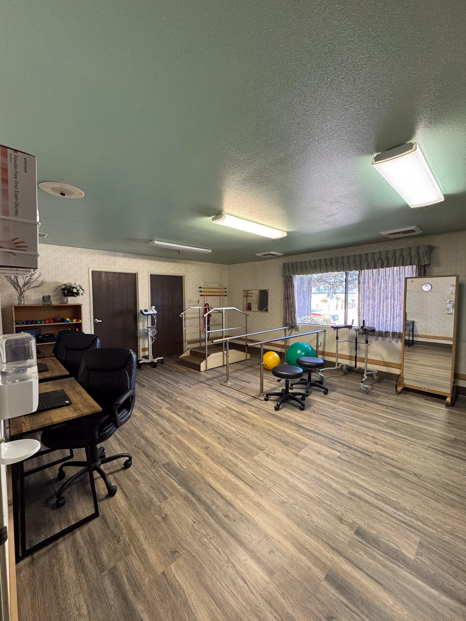 A bright rehab/exercise room with parallel bars, therapy balls, rolling stools, desks and chairs on wood flooring.