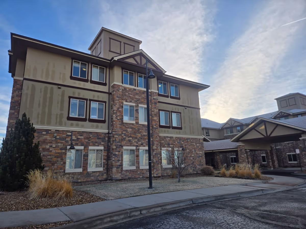 Three-story assisted living building with stone and stucco facade and a covered entrance under a cloudy sky.
