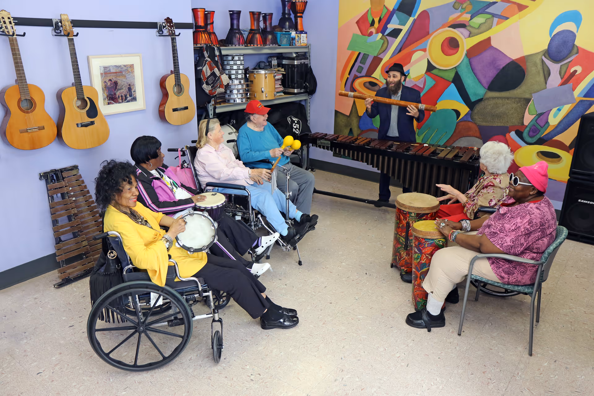 A group of elderly people, some in wheelchairs, participating in a music therapy session in a room decorated with guitars on the wall and colorful percussion instruments. A man is playing a large wooden flute in front of a vibrant mural featuring abstract musical instruments.