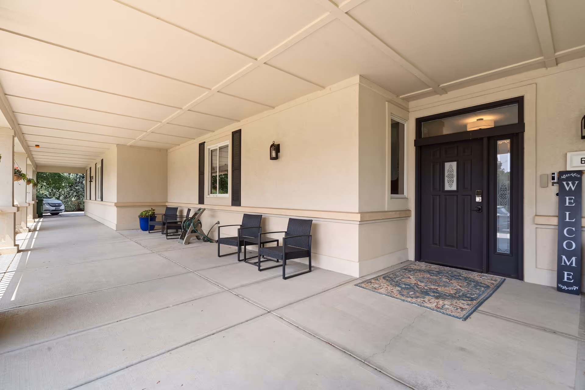 Covered outdoor walkway with chairs, a dark entrance door, and a welcome sign.