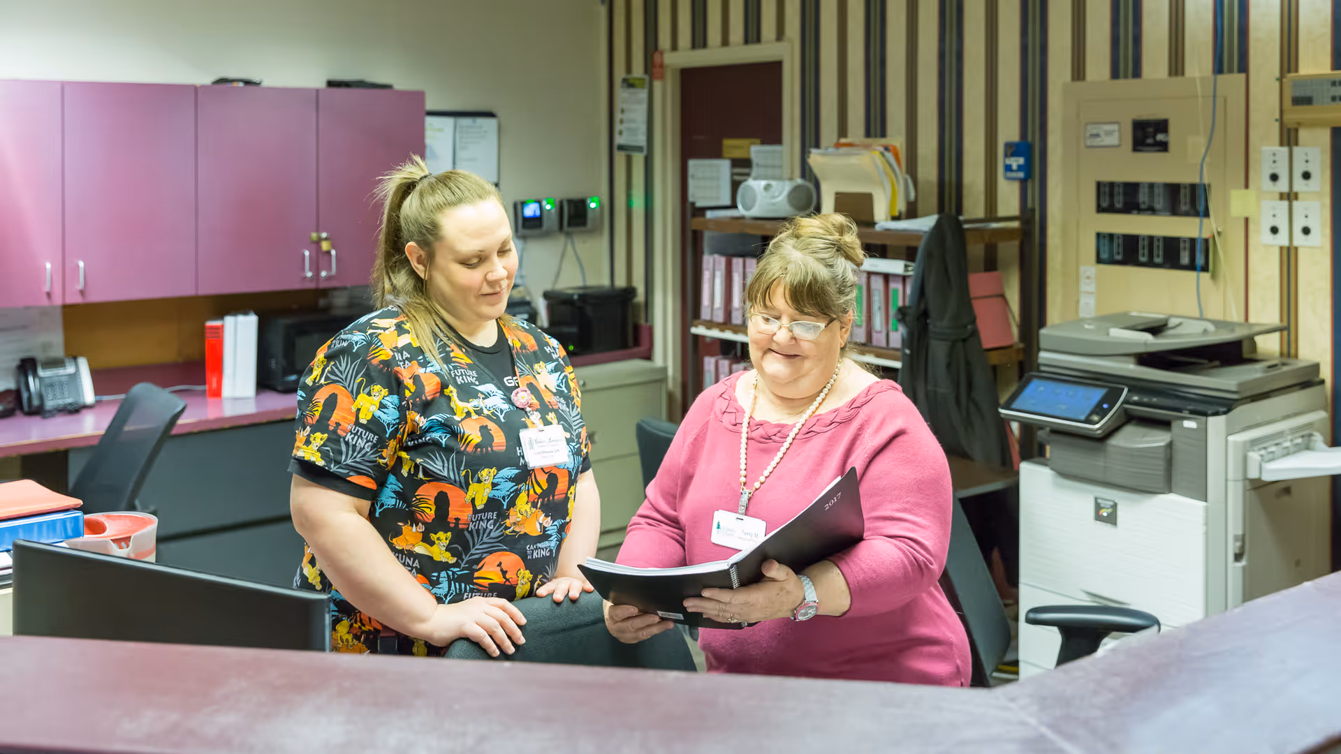 Two women standing behind a reception desk in an office setting. One woman is wearing a colorful scrub top with animal prints, and the other woman is wearing a pink sweater and glasses, holding and looking at a black folder. The background shows office equipment, filing shelves, and cabinets.