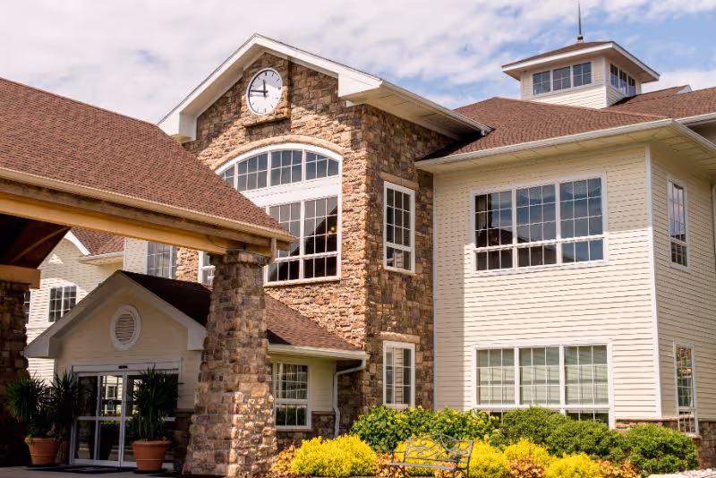 Entrance and facade of The Heritage Of Green Hills senior living building with stone accents, large windows, a clock above, and landscaped greenery.