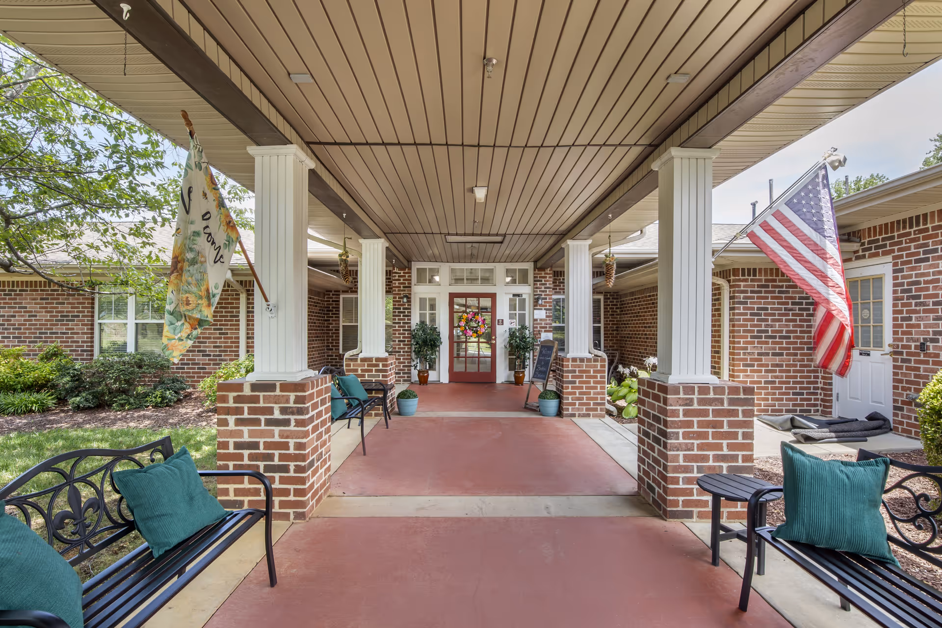Covered entrance area of a brick building with white columns and a red door decorated with a colorful wreath. There are benches with green cushions on either side, potted plants, and two flags hanging—one floral and one American flag. The area is surrounded by greenery and has a red concrete floor.