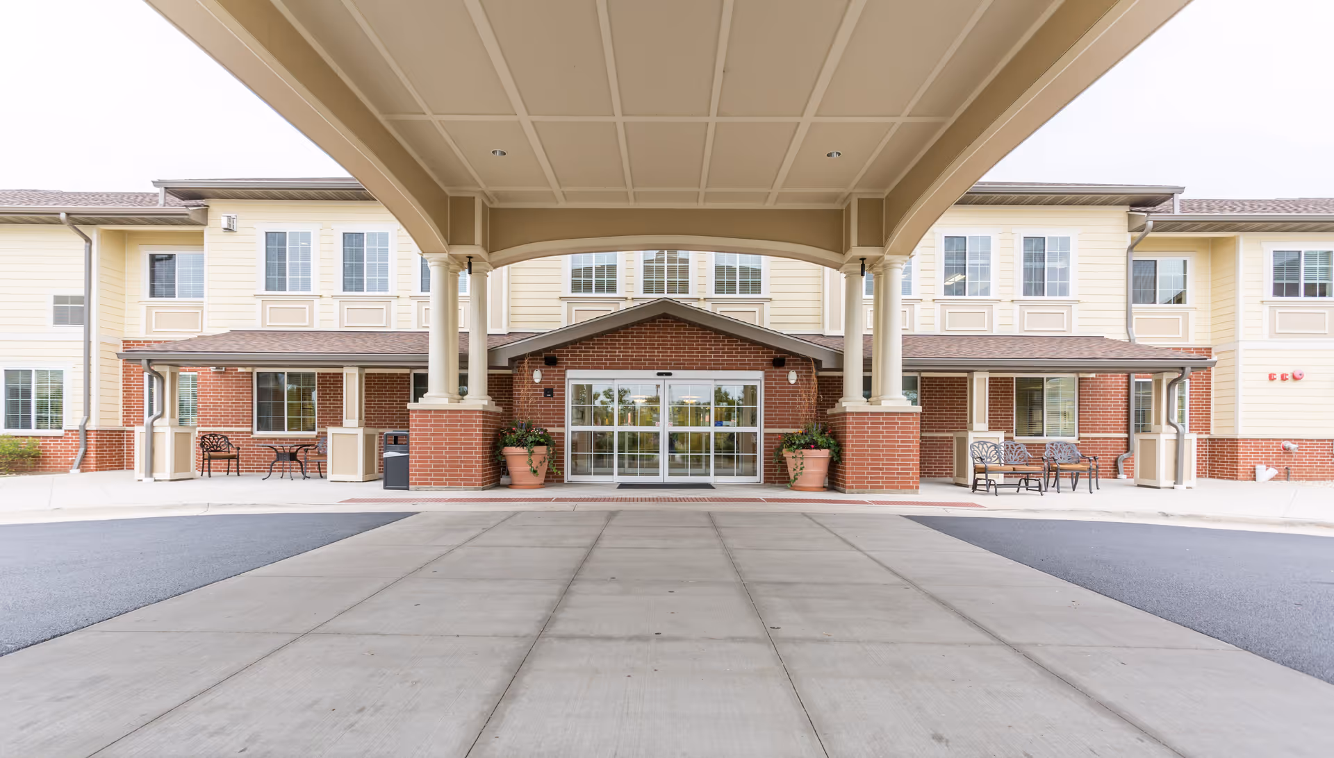 Front exterior view of Three Oaks Assisted Living and Memory Care building with a covered entrance supported by columns, large glass doors, brick and beige siding, and outdoor seating areas on both sides.