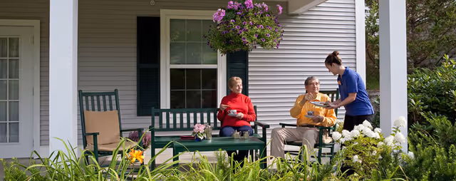 Three people and a caregiver chat on a shaded terrace porch outside a senior living facility.