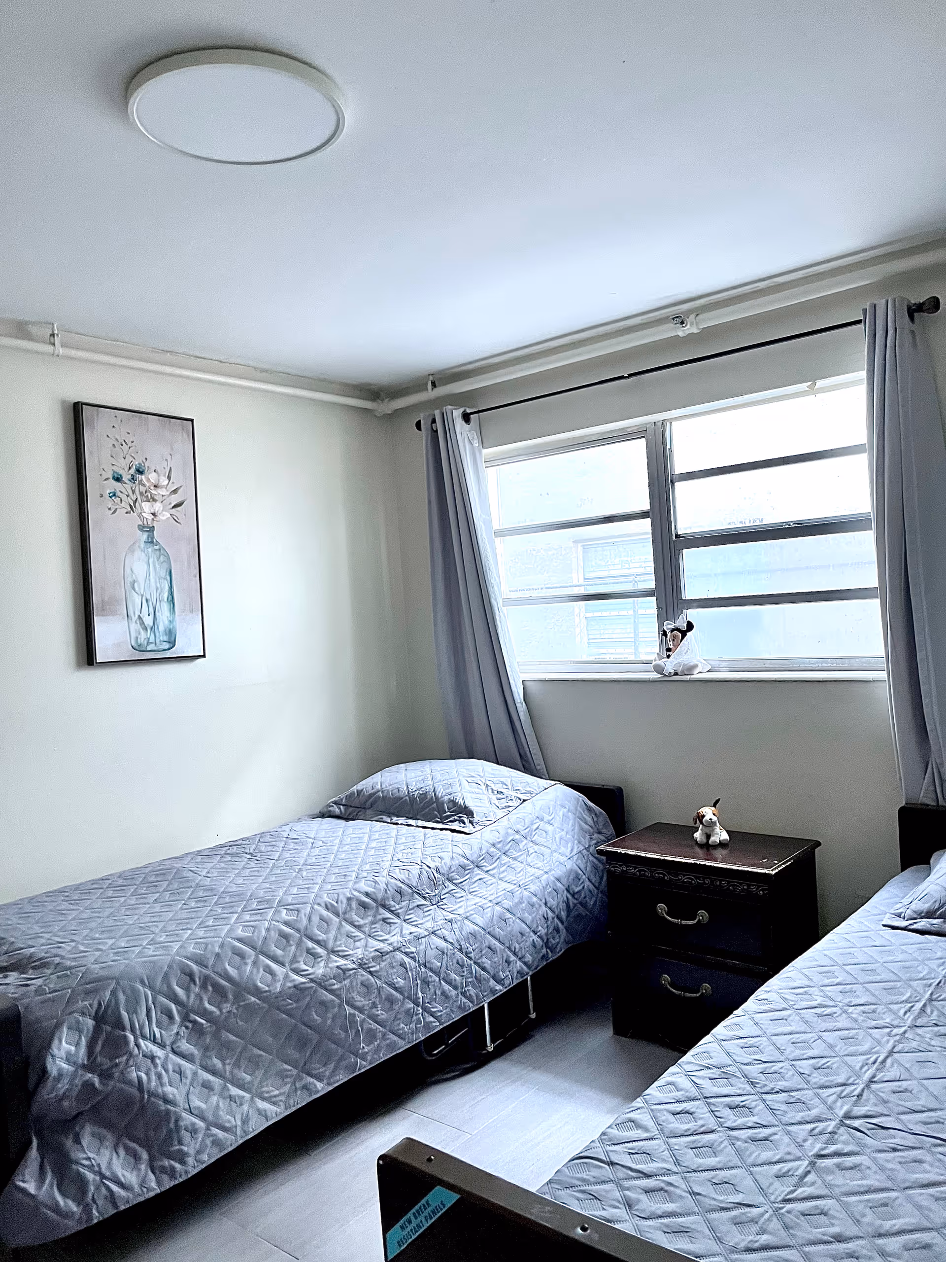 A small bedroom with two single beds covered in gray quilted bedspreads. Between the beds is a dark wooden nightstand with two drawers, topped with a small stuffed animal. A window with gray curtains lets in natural light. A framed picture of a vase with flowers hangs on the wall above one bed.