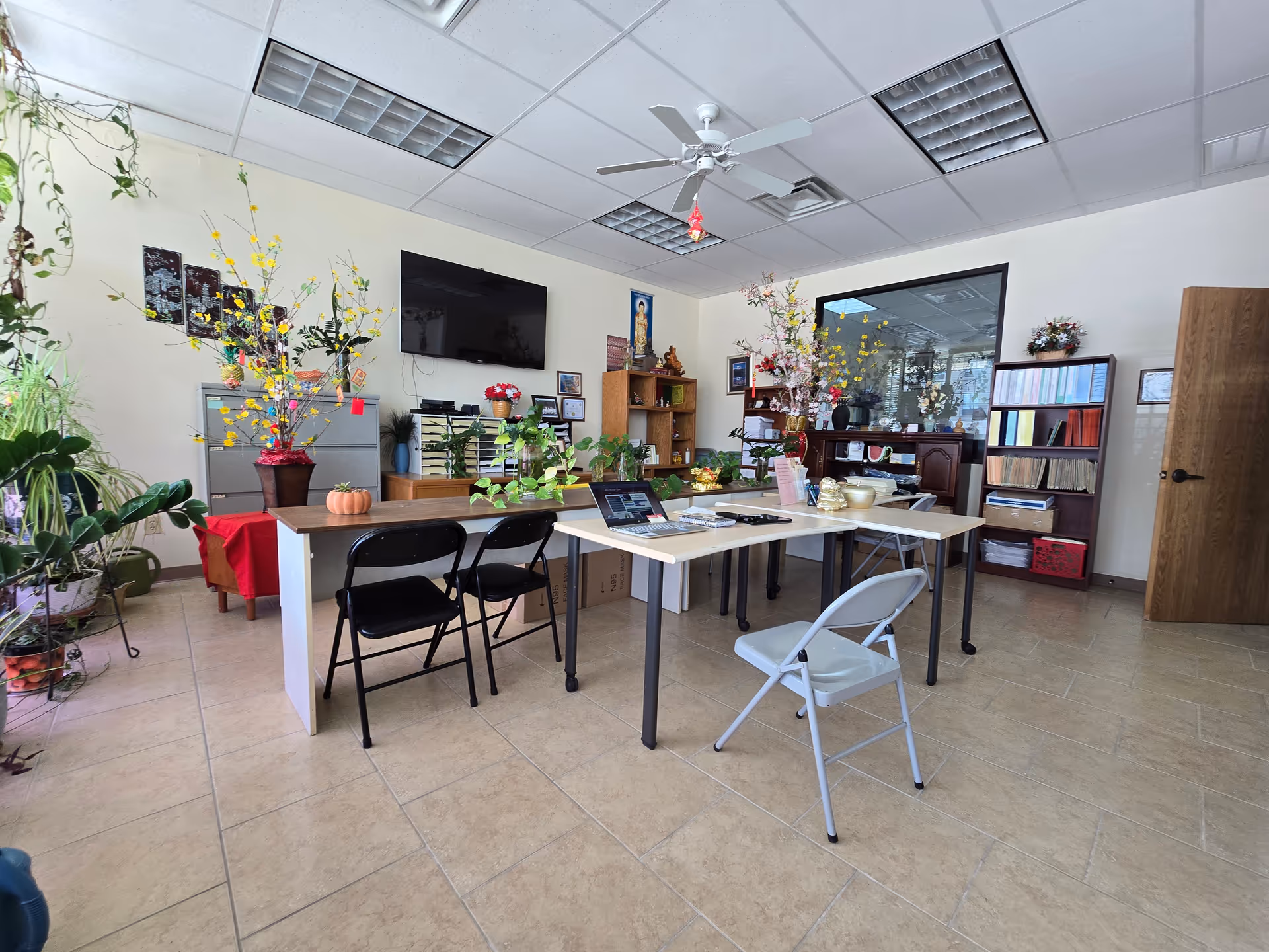 Interior common room with tables, chairs, plants, shelves, and a wall-mounted TV.