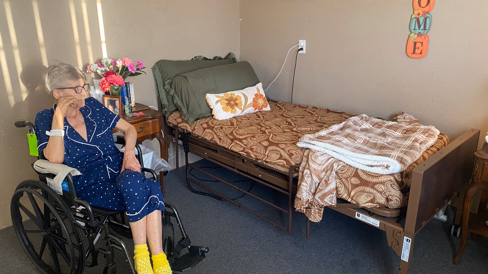 Senior woman in a wheelchair sits beside a decorated bed in a cozy bedroom.